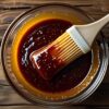 Glass bowl of golden barbecue glaze with a basting brush on a rustic wood surface