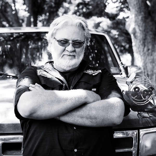 Portrait of Myron Mixon leaning on a car, arms crossed, staring confidently at the camera