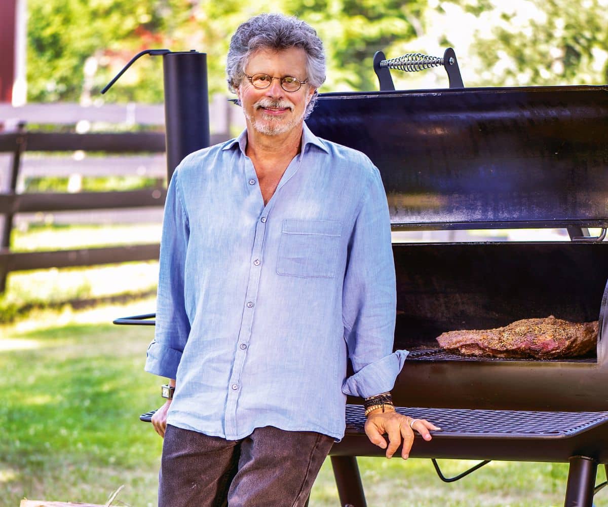 Steven Raichlen standing in front of an open smoker with a brisket inside, looking toward the camera